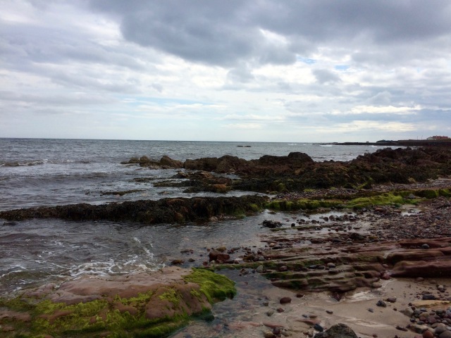 A landscape showing the shoreline at Barns Ness. There are red rocks in the foreground, the sea in the background, and stratus clouds overhead.